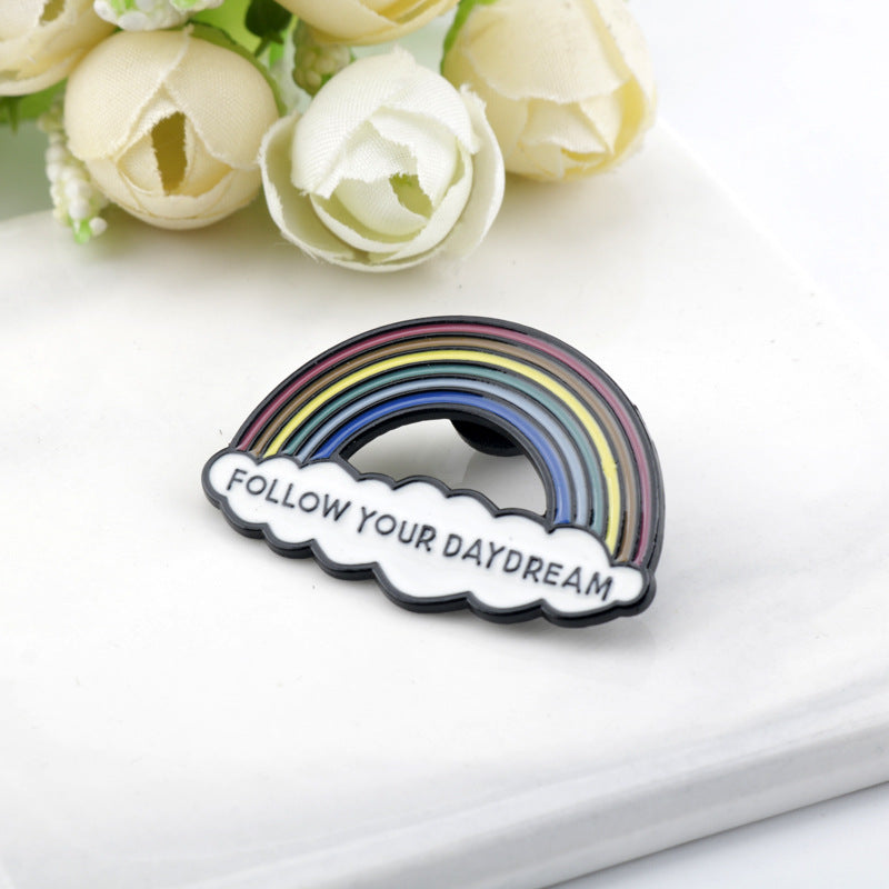 Rainbow bridge metal brooch reading “FOLLOW YOUR DAYDREAM” with cloud accents