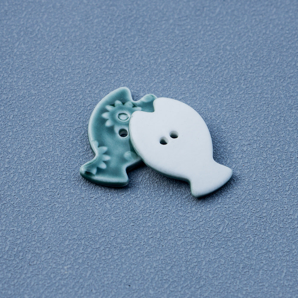 Green fish-shaped ceramic button with 2 holes and raised flower design, side view.