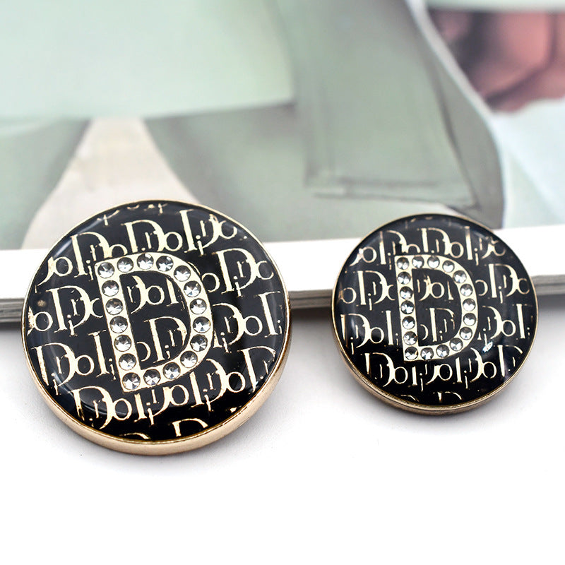 Black round button with rhinestone letter 'D' and gold-toned border