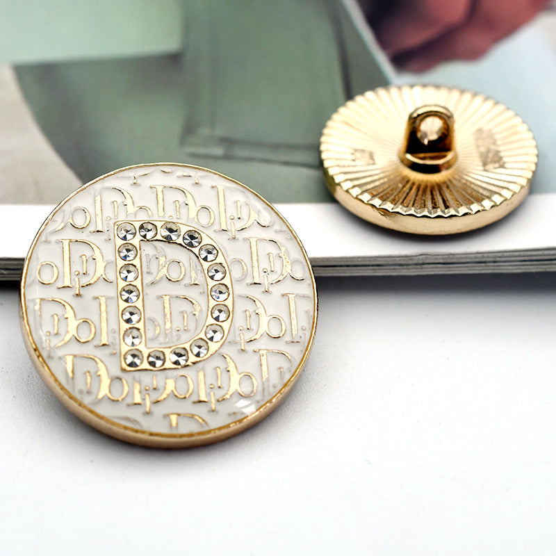 White round button with rhinestone letter 'D' on ornate pattern, gold-toned rim