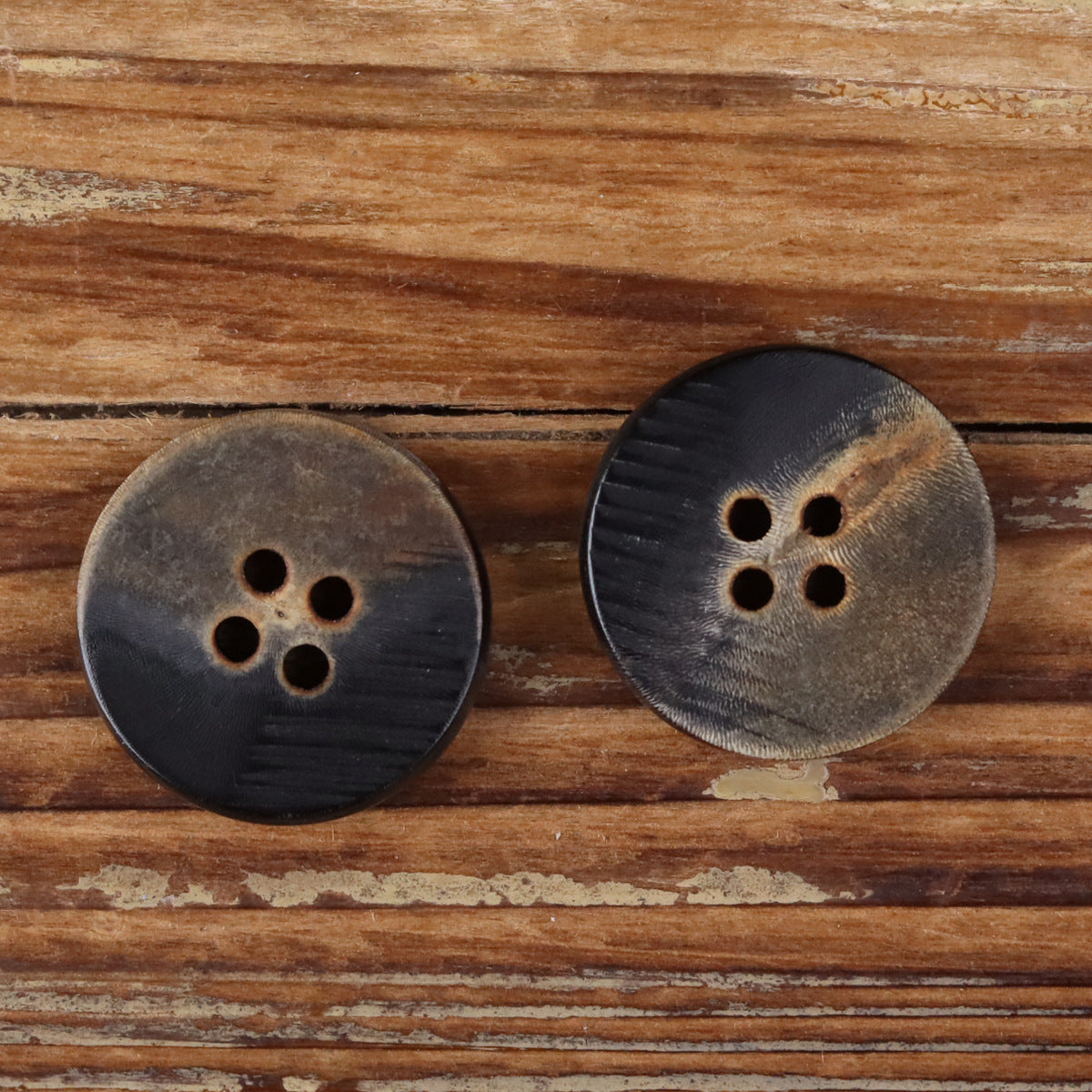 Two dark brown-black 4-hole flat buttons with horn-grain texture and scorched edge