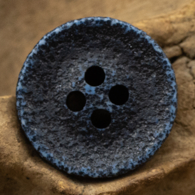 Black distressed resin button, four round holes, mottled textured surface.