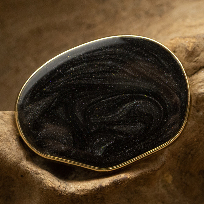 Black irregular oval pearl-look glass button with glossy swirl pattern and gold edge.