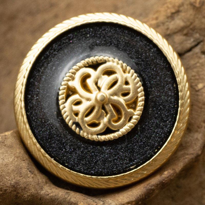 Pearl-glaze shank button in black with ornate gold filigree center and ridged rim