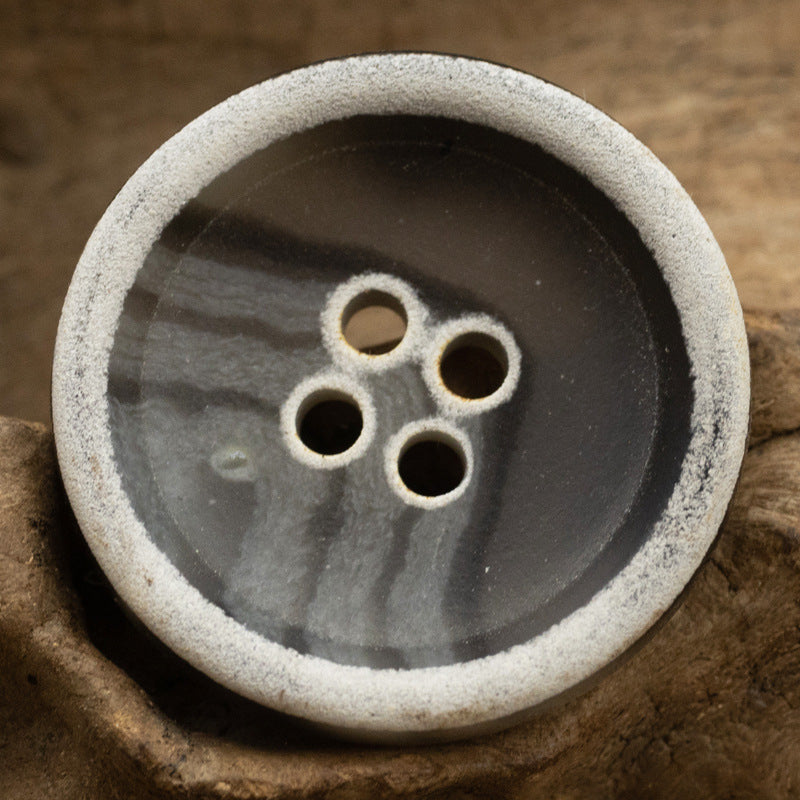 Dark gray-black button, textured face with 4 holes, light burnt-edge underside