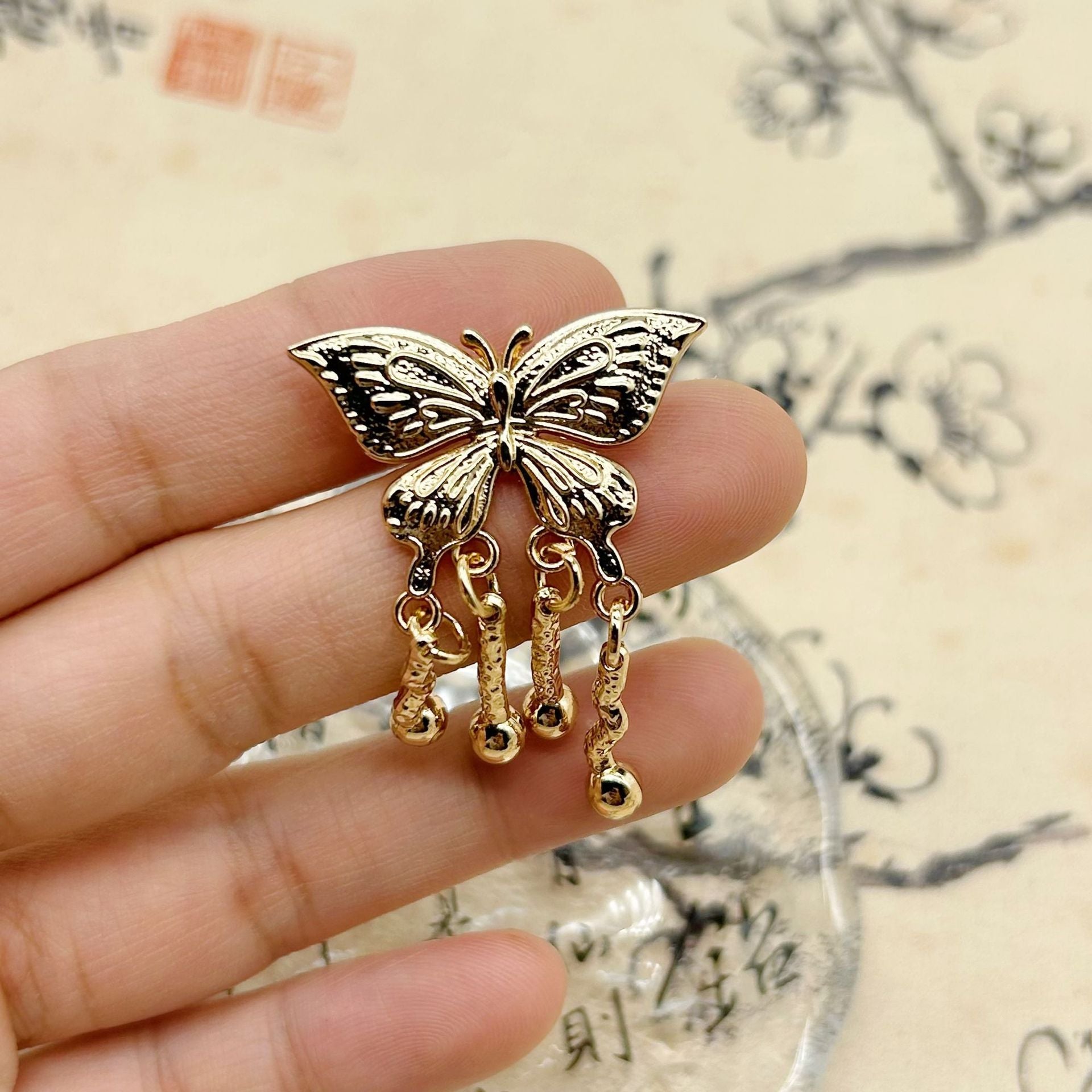 Golden butterfly tassel button shown on fingertip, ornate metal filigree and beads