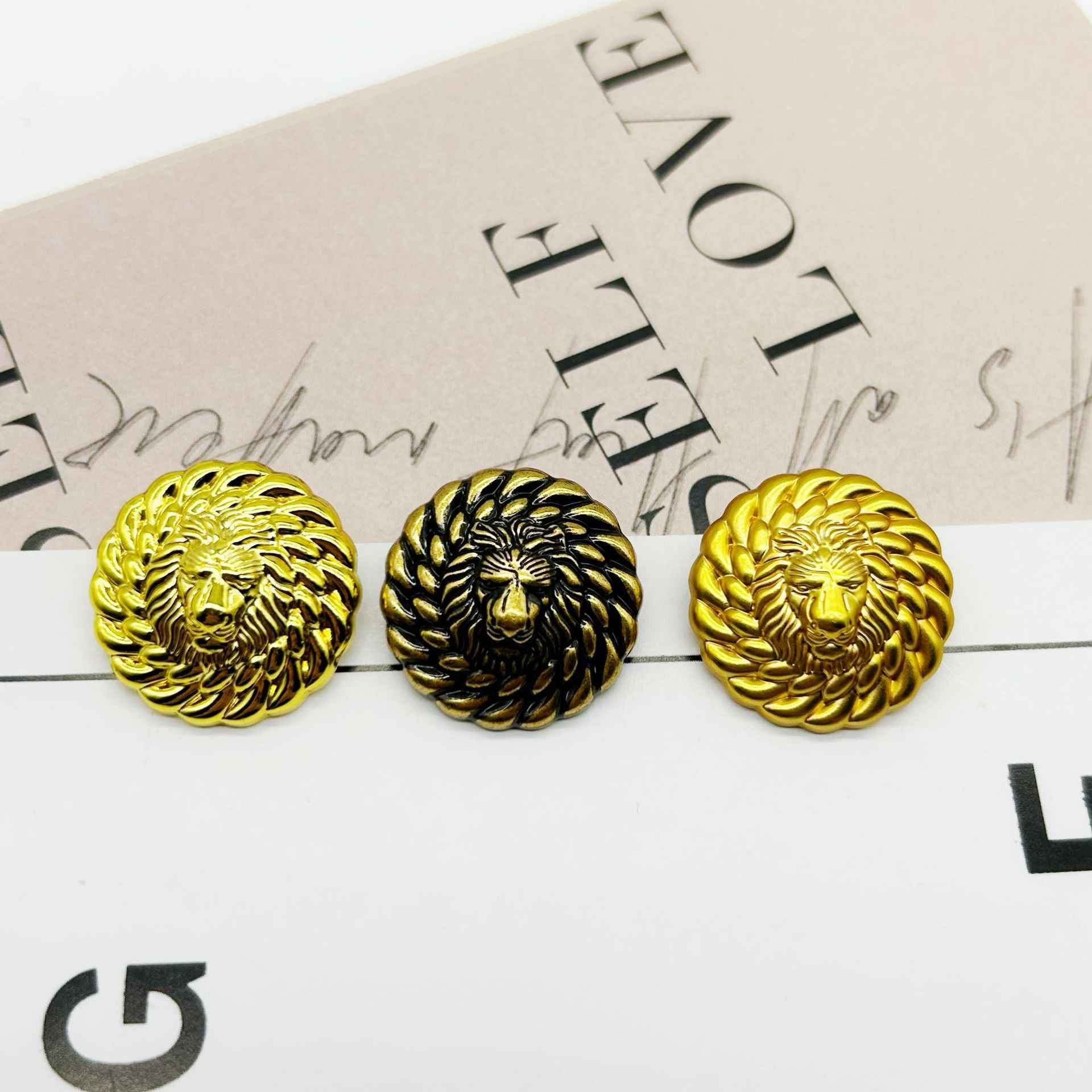 Three round lion head buttons in gold tones, one darker finish, embossed faces