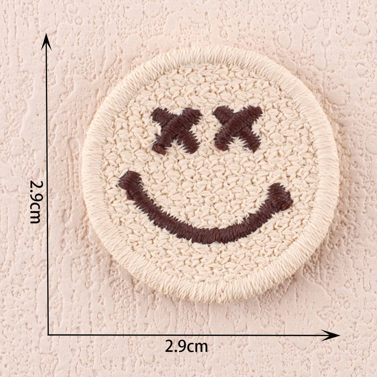 Cream fabric patch with dark brown smiley face stitches and X-shaped eyes, 2.9cm.