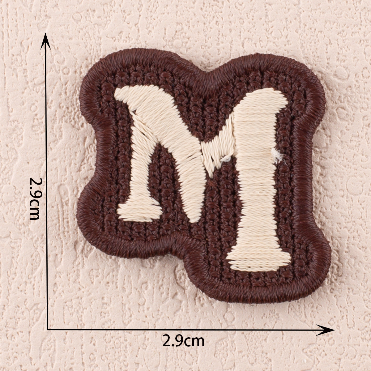 Dark brown fabric patch with white letter M, 2.9cm x 2.9cm stitched shape.