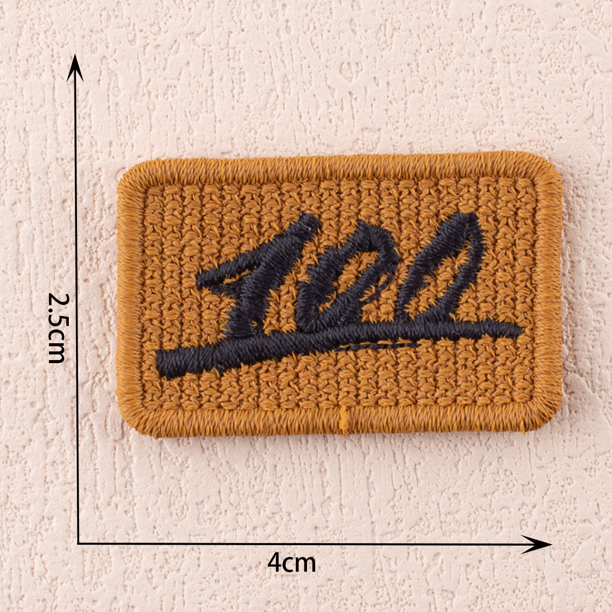 Brauner Stoffpatch mit dunkelblauer bestickter Buchstaben-/Ziffernform, ca. 4×2,5 cm