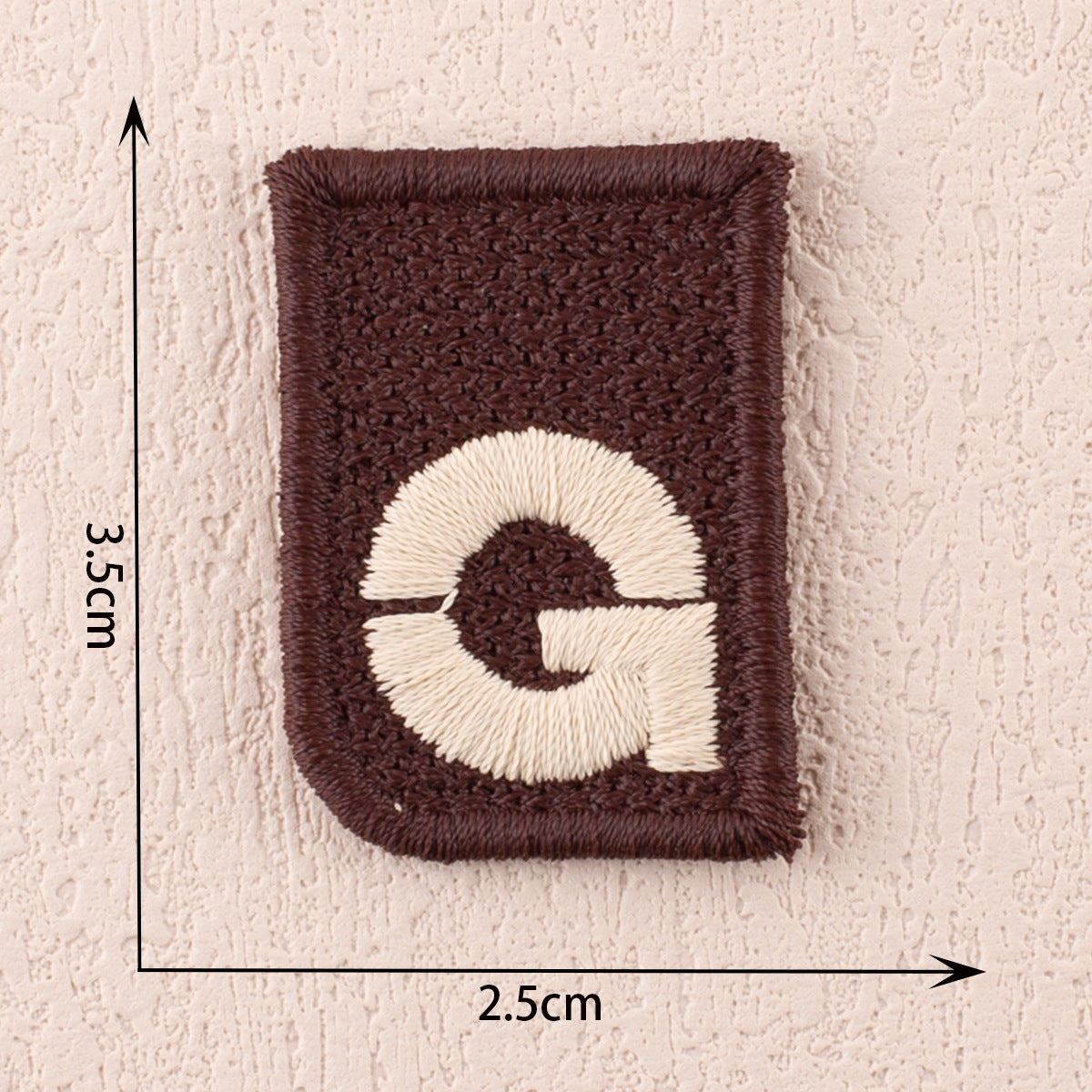 Dark brown fabric patch with white letter G and a curved underline, 2.5cm x 3.5cm.
